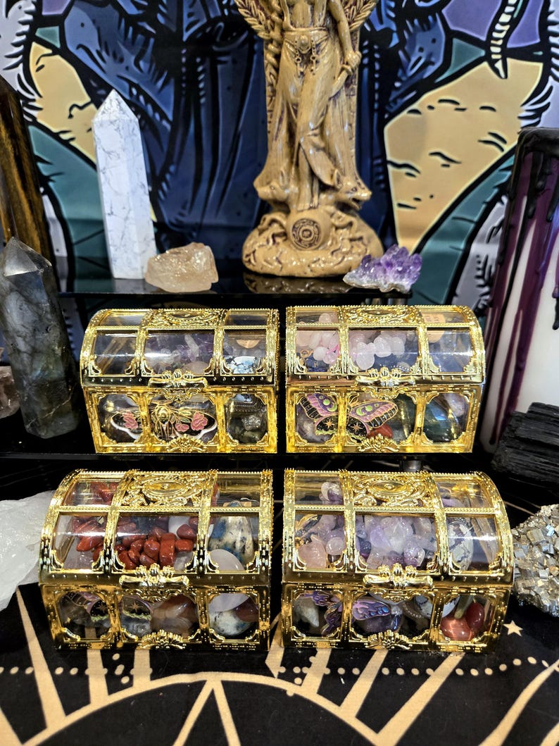 May include: Four small, ornate gold treasure chests with clear tops, each filled with colorful gemstones. The chests are arranged on a black surface, with a statue and crystals in the background. The chests have decorative gold accents.