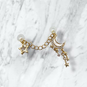 May include: Gold-toned ear cuff featuring a star and a crescent moon charm, connected by a delicate chain. The moon is embellished with white enamel and sparkling crystals, with dangling star accents. The ear cuff is designed to be worn on one ear.
