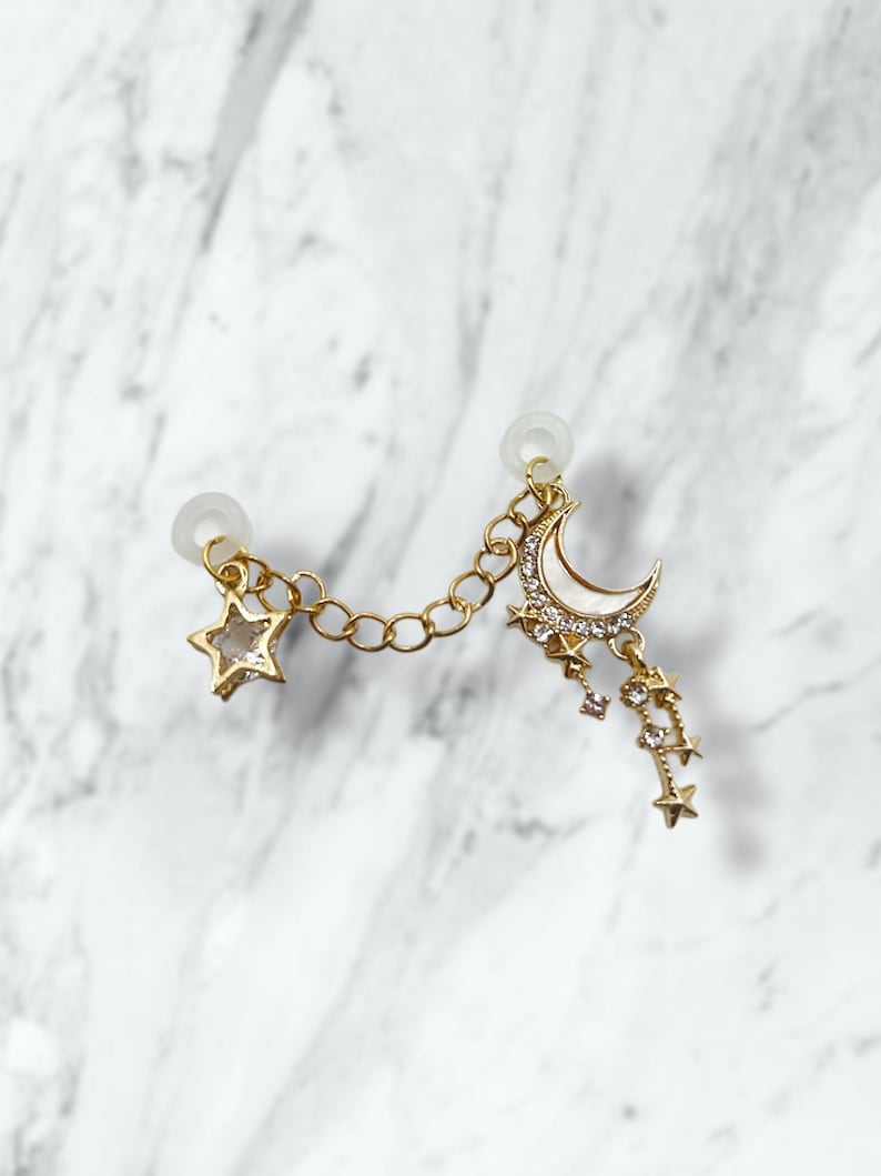 May include: Gold-toned ear cuff featuring a star and a crescent moon charm, connected by a delicate chain. The moon is embellished with white enamel and sparkling crystals, with dangling star accents. The ear cuff is designed to be worn on one ear.