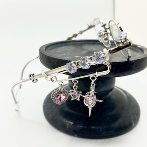 May include: Silver-toned eyeglasses with clear lenses, adorned with sparkling purple and clear gemstones. The frames feature dangling charms, including a pink heart, a star, and a ballerina. The glasses rest on a dark, round pedestal.