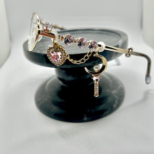 May include: Gold-toned glasses featuring a decorative side arm adorned with clear round crystals, a dangling pink heart-shaped charm, a gold chain, and a crescent moon charm with a small pearl and a vertical crystal-encrusted bar pendant.