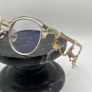 May include: Gold-rimmed eyeglasses with clear lenses. The temples feature a decorative design and are adorned with a charm of a star, moon, and smaller stars. The glasses are displayed on a dark gray marble stand.