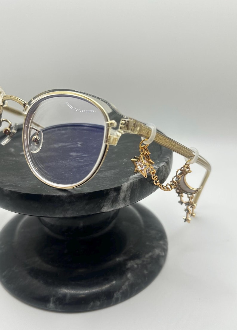 May include: Gold-rimmed eyeglasses with clear lenses. The temples feature a decorative design and are adorned with a charm of a star, moon, and smaller stars. The glasses are displayed on a dark gray marble stand.
