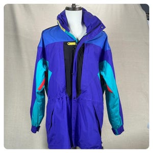 May include: A vintage ski jacket in a colour-blocked design, featuring shades of blue, purple, teal, and red. The jacket has a high collar, drawstring waist, and a black front zip. The sleeves have teal and red accents. The jacket is displayed on a mannequin.