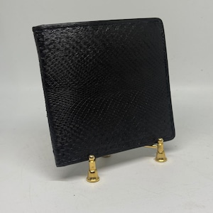 Könnte beinhalten: Eine schwarze, strukturierte Lederbrieftasche. Die Brieftasche ist quadratisch und scheint aus einem Material mit schuppenartigem Muster zu bestehen. Die Brieftasche wird auf einem kleinen, goldfarbenen Ständer mit drei Beinen präsentiert.