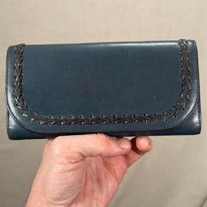 Puede incluir: Una cartera de cuero color verde azulado con un diseño de borde trenzado. La cartera tiene una solapa curva y la marca "Chloe" es visible. La cartera se sostiene en una mano.