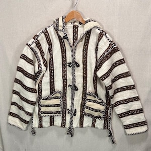 May include: A hooded jacket with a cream and brown striped pattern. The jacket has two front pockets, toggle closures, and decorative stitching along the edges. The jacket is hanging on a wooden hanger.