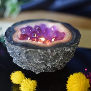 Peut inclure: Un photophore décoratif en géode d'améthyste. La géode est de couleur gris foncé avec une texture rugueuse et un intérieur lisse de couleur claire. L'intérieur est rempli de cristaux violets et d'une lueur chaude.