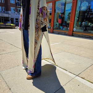 May include: A long, cream-colored kimono with a floral pattern in purple, brown, and orange. The kimono is worn over a black top and blue jeans. The outfit is completed with woven shoes.