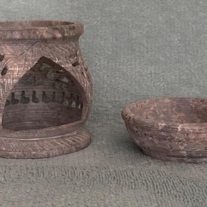 May include: A carved stone oil warmer and a small bowl. The oil warmer has a cylindrical shape with intricate floral cutouts and a triangular opening. The bowl is round and has a textured surface. Both items are in shades of brown.