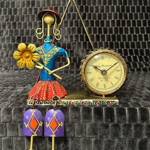 May include: A whimsical metal figurine clock featuring a woman holding a yellow flower, with a clock face that reads "Village Clockworks, Birmingham est. 1798". The woman wears a blue top, red skirt, and a red hat. The clock has two purple bell-shaped feet.
