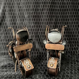 May include: Two decorative miniature motorcycle sculptures. The motorcycles are made of copper-colored metal with brown accents. Each motorcycle has a round headlight, a seat, and saddlebags with white floral designs. The background is a dark gray textured fabric.