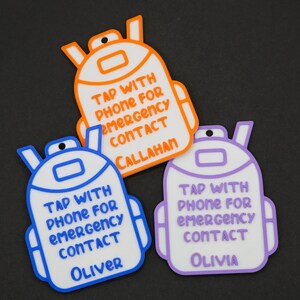 May include: Three colorful backpack-shaped tags. Each tag has a white background with a different colored outline: orange, blue, and purple. Text on each tag reads "TAP WITH PHONE FOR EMERGENCY CONTACT" followed by a name: Callahan, Oliver, and Olivia.