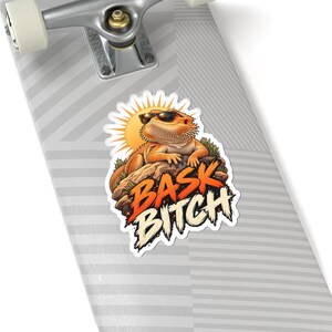 May include: A sticker on a skateboard deck featuring an orange bearded dragon wearing black sunglasses, resting on a rock under a yellow sun. The text BASK BITCH is printed in bold orange and white letters below the lizard on the sticker.