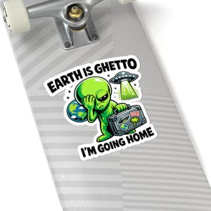 May include: A sticker featuring a green alien with a sad expression, holding a suitcase and a globe. The text "EARTH IS GHETTO" and "I'M GOING HOME" are displayed. A UFO is also depicted.