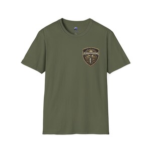 Puede incluir: Camiseta verde oliva con cuello redondo. Presenta un parche en forma de escudo en el pecho con el texto "ATDAGGERSORMAN" sobre un diseño de daga. El parche tiene una combinación de colores marrón y dorado.