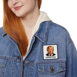 Könnte beinhalten: Blaue Jeansjacke mit einem Aufnäher mit einem Porträt mit Schnurrbart und dem Wort "REDACTED" in weißer Schrift. Die Jacke hat eine Knopfleiste, Taschen und einen hellfarbigen Kapuzenpullover darunter.