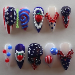 May include: A set of ten press-on nails featuring red, white, and blue designs with 3D elements including strawberries, ice cream cones, bows, and stars. The text @lumpy.nails is visible on the first nail. The nails have a pointed stiletto shape.