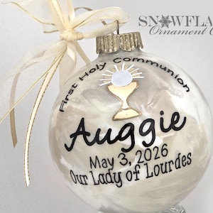 May include: A clear glass spherical ornament filled with white material, featuring a gold chalice and host graphic. Black text reads First Holy Communion, Auggie, May 3, 2026, and Our Lady of Lourdes. A cream-colored ribbon is tied to the silver metal cap.