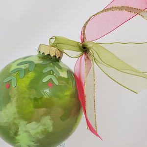May include: A green glass Christmas ornament with a decorative leaf and berry design. The ornament is adorned with a sheer pink and green ribbon with gold trim. The ornament is round and has a gold-colored metal cap.