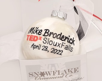 May include: A round white glass ornament with a silver cap and a sheer ribbon. The text reads Mike Broderick TEDx SiouxFalls April 28, 2022 in black and red lettering. The ornament is nestled in a white box labeled Snowflake Ornament Co.