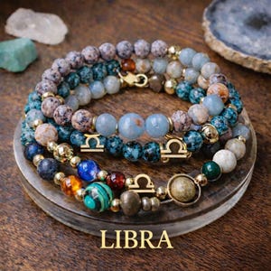 May include: A multi-layered beaded bracelet featuring a mix of speckled, blue, and earth-toned stones, interspersed with gold-colored metal beads and gold-colored Libra zodiac symbol charms. The word LIBRA is printed in gold at the bottom.