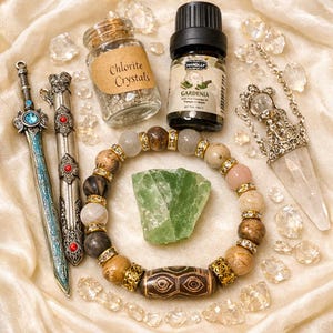 May include: An assortment of spiritual items including a beaded bracelet, a green crystal, a bottle of essential oil, a crystal pendulum, and two decorative swords. A small jar of Chlorite Crystals is also visible.