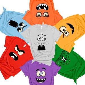 May include: A selection of brightly coloured t-shirts, each with a different cartoon face design. The shirts are in red, orange, yellow, green, blue, grey, and purple. The faces display a range of expressions, including anger, surprise, and happiness.