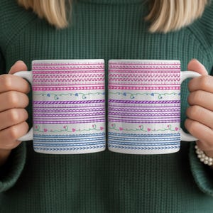 Puede incluir: Dos tazas de cerámica blanca con un diseño colorido y estampado. El diseño presenta bandas horizontales rosas, moradas, azules y verdes con elementos decorativos como corazones y zigzags. Las tazas son sostenidas por una persona.