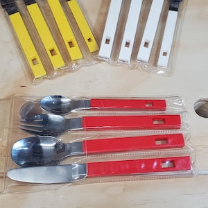Puede incluir: Una colección de juegos de cubiertos empaquetados. Los juegos incluyen tenedores, cucharas y cuchillos con cabezas de metal negro y mangos amarillos, blancos y rojos. Los utensilios están en envases de plástico transparente.