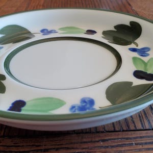 May include: White ceramic saucer with a green rim and a central recessed area. The saucer is decorated with green leaves, blue flowers, and dark blue accents. The saucer is on a wooden surface.