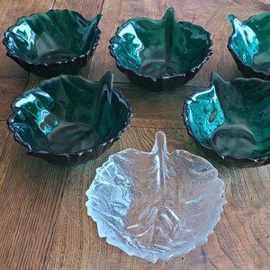 Op de afbeelding: Een set van zes decoratieve kommen in de vorm van een blad. Vijf zijn diep smaragdgroen en één is helder glas. De kommen zijn gerangschikt op een houten oppervlak, waarbij hun unieke ontwerp en kleurvariaties worden getoond.