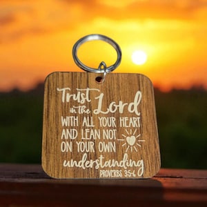 Può includere: Portachiavi in legno con anello argentato. Il portachiavi presenta la scritta "Trust in the Lord" e un versetto biblico. Lo sfondo mostra un tramonto.