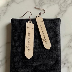 Puede incluir: Un par de pendientes de madera con la frase "I am enough" grabada en cursiva. Los pendientes son de color marrón claro y cuelgan de ganchos plateados. Se exhiben sobre una superficie negra texturizada.