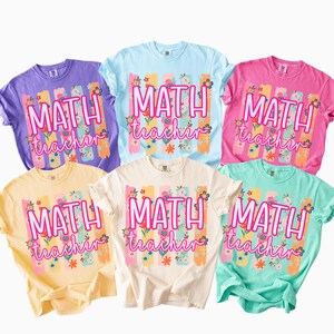 May include: Five short-sleeved crew neck t-shirts in purple, light blue, pink, yellow, and mint green. Each shirt features a central graphic with vertical brushstrokes in pastel colors, floral accents, and the text "MATH teacher" in pink.
