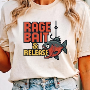 May include: Beige t-shirt with a pixelated design featuring the words "RAGE BAIT & RELEASE" in red and yellow block letters. A pixelated fish with red and black scales is hooked by a fishing hook.