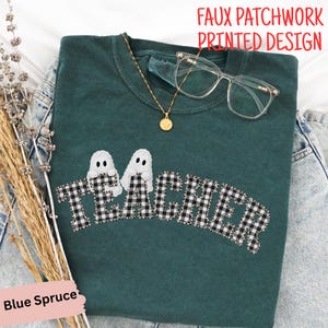May include: A blue spruce colored t-shirt featuring the word TEACHER in a black and white buffalo plaid faux patchwork printed design. Two white ghost figures are positioned above the letters. A gold necklace and clear glasses are included.