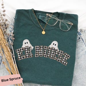 May include: A dark green crewneck t-shirt featuring the text ER NURSE in a black and white buffalo plaid pattern. Two white ghost illustrations are positioned above the text. A gold chain necklace and clear glasses rest on the shirt.
