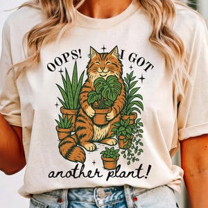 May include: Beige t-shirt featuring a smiling orange tabby cat holding a potted plant, surrounded by other potted plants. The text reads "OOPS! I GOT another plant!" in black, whimsical lettering. The design has a vintage, illustrated style.