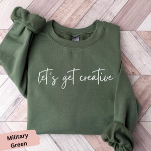 May include: An olive green sweatshirt with the phrase "let's get creative" in white script. The sweatshirt has a crew neck and long sleeves. The bottom left corner of the image has a pink label that says "Military Green".