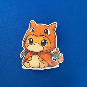 May include: A sticker featuring a cartoon character in an orange hooded outfit with a cape and small green wings. The character has large eyes and a light orange body. The sticker is on a blue background.