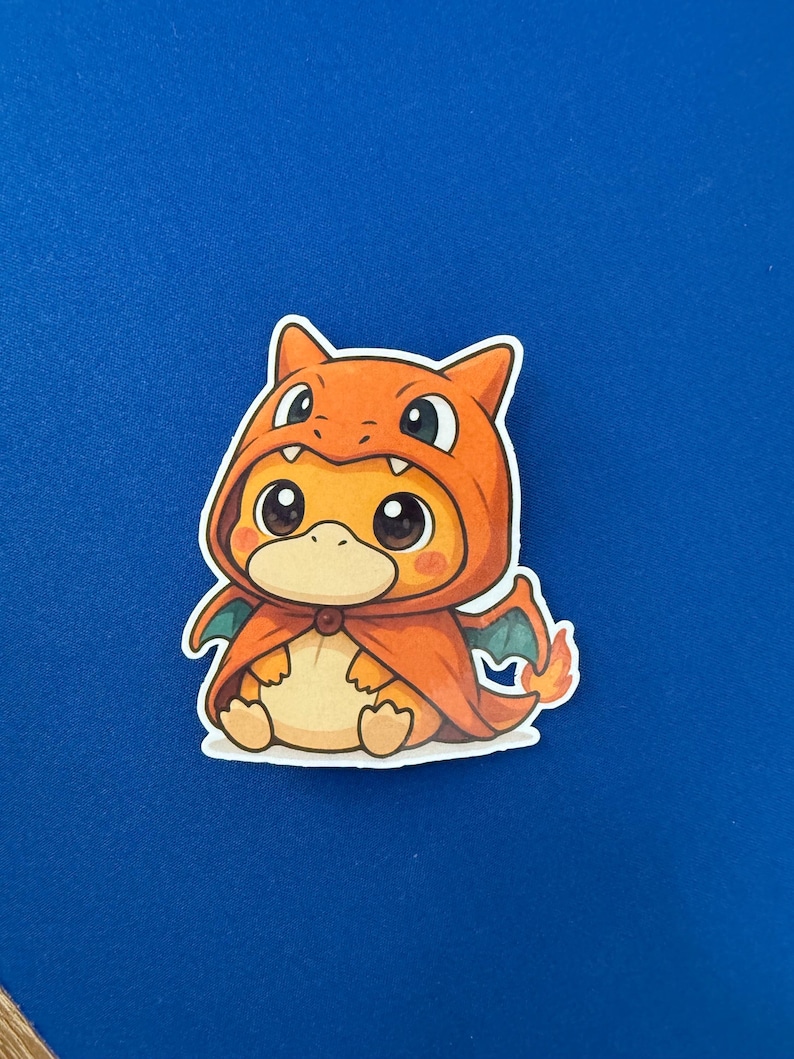 May include: A sticker featuring a cartoon character in an orange hooded outfit with a cape and small green wings. The character has large eyes and a light orange body. The sticker is on a blue background.