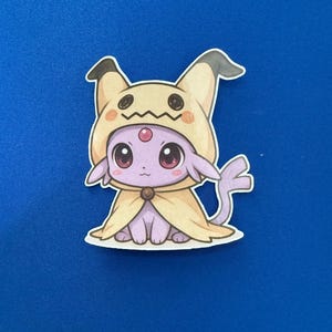 May include: A sticker featuring a cartoon character in a Mimikyu costume. The character is purple with large, pink eyes and a yellow hooded cloak. The background is a solid blue.