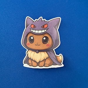 May include: A sticker featuring a cartoon character in a purple hooded cloak with red eyes and a smiling mouth. The character has brown fur, large eyes, and a cream-colored chest. The sticker is on a blue background.