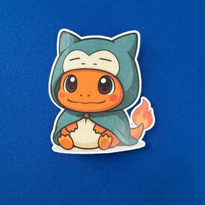 May include: A sticker featuring a cartoon Charmander character dressed as Snorlax. The Charmander is orange with a white belly, wearing a teal cape and hood. A small flame is visible on its tail. The sticker is on a blue background.