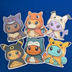 May include: Six colorful cartoon character stickers on a blue background. The stickers feature various creatures dressed in hooded costumes, including a purple Gengar, orange Charmander, and yellow Pikachu. The stickers are outlined in white.