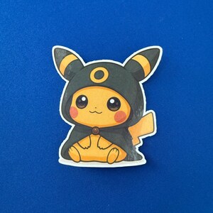 May include: A die-cut sticker featuring a yellow Pikachu character wearing a black hooded cloak with yellow accents. The character has rosy cheeks and is sitting down. The background is a solid blue.