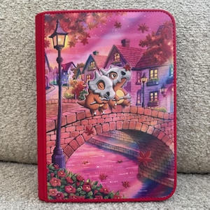 May include: A rectangular zippered binder featuring a vibrant illustration of two Cubone characters sitting on a brick bridge over a pink river. The scene includes a street lamp, houses, and autumn leaves in shades of pink, purple, and orange.