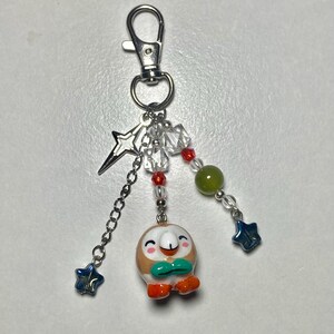 May include: A silver-toned metal keychain featuring a small, round, brown and white bird-like character charm with orange feet and a green bowtie. The charm is accented with clear, red, and green beads, a silver star charm, and two blue star beads.