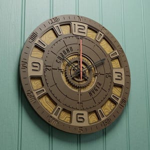 Könnte beinhalten: Eine kreisförmige Wanduhr mit geschichtetem Design, bestehend aus einem dunkelbraunen Rahmen und einem hellen, holzfarbenen Hintergrund mit topografischen Linien. Das Zifferblatt zeigt den Text CHRONO und NVGTR, große Ziffern, ein zahnradartiges Zentrum und einen roten Sekundenzeiger.